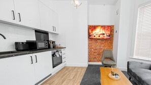 Een moderne keuken met witte kasten, een magnetron en een koffieapparaat. Er is een metselwerk open haard met een televisie erboven, een grijze fauteuil en een grijze bank.