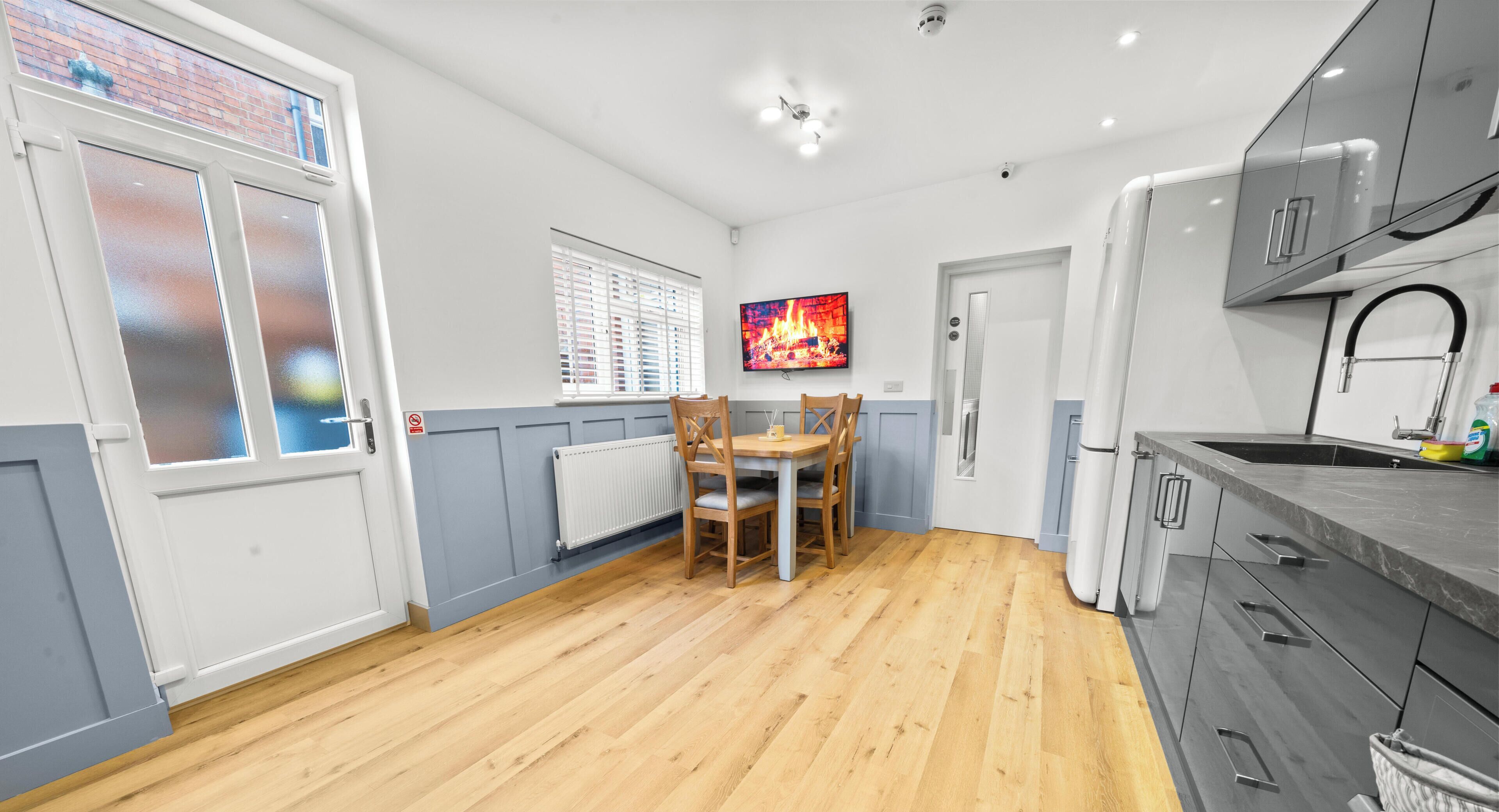 A room with a dining table, chairs, a television mounted on the wall, and a kitchen area with cabinets and a refrigerator.