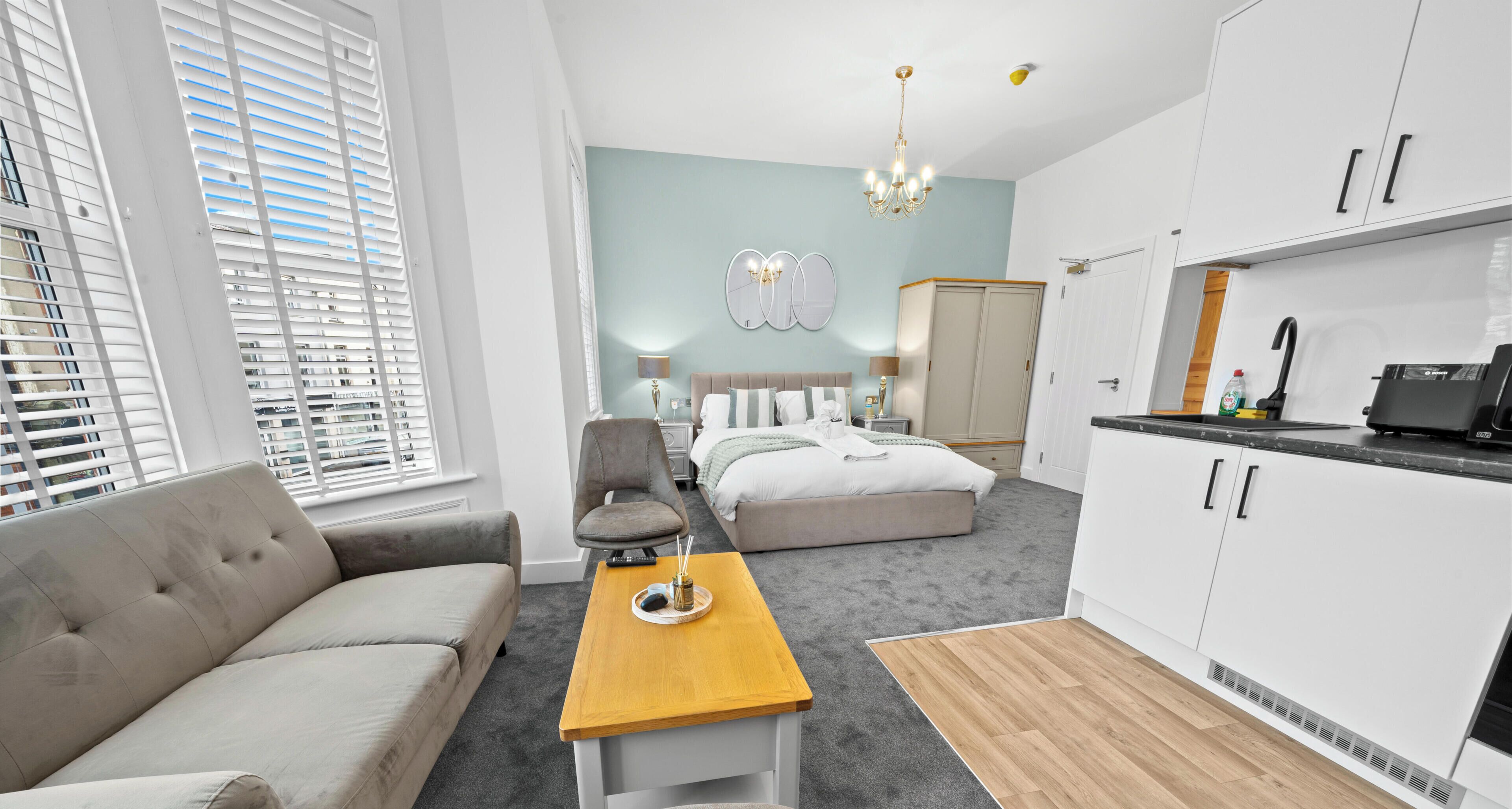 A modern bedroom with a sofa, a wooden table, a bed, a chair, and a kitchenette.