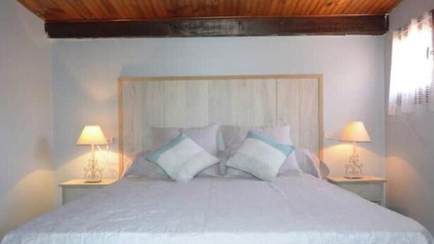 Un dormitorio con techo de madera, una cama con ropa de cama blanca, dos lámparas de noche y una mesita de noche con lámpara.
