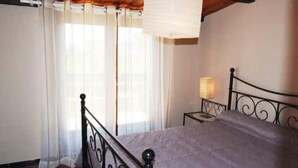 Un dormitorio con cama, mesita de noche, lámpara, ventana con cortinas traslúcidas y techo de madera.