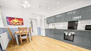 A modern kitchen with grey cabinets, a built-in oven, a microwave, and a dining area with a wooden table and chairs. - Burnham-on-sea | Warm, Bright Ensuite Room | Ideal for Couple | Contractors Stay (Somerset)