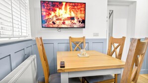 A room with a television displaying a fireplace, a table with chairs, and a window with blinds. - Burnham-on-sea | Warm, Bright Ensuite Room | Ideal for Couple | Contractors Stay (Somerset)