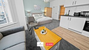 A modern bedroom with a bed, bedside tables, and a chair. There is a kitchenette with a microwave and oven. The room has a light blue wall and a chandelier. - Burnham-on-sea | Luxury | Families | Large Groups (Somerset)