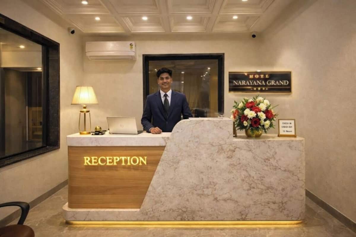 Een hotel receptiebalie met een bord waarop 'Hotel Narayana Grand' staat, een laptop, een lamp en een bloemstuk.