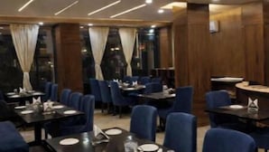 A restaurant with dark wooden paneling, blue upholstered chairs, and tables set with white plates and glasses. - Narayana Grand Hotel (New Delhi)