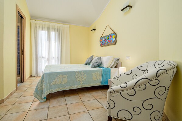 Un dormitorio con cama, una silla, ventana con cortinas, un adorno de pared y una puerta.