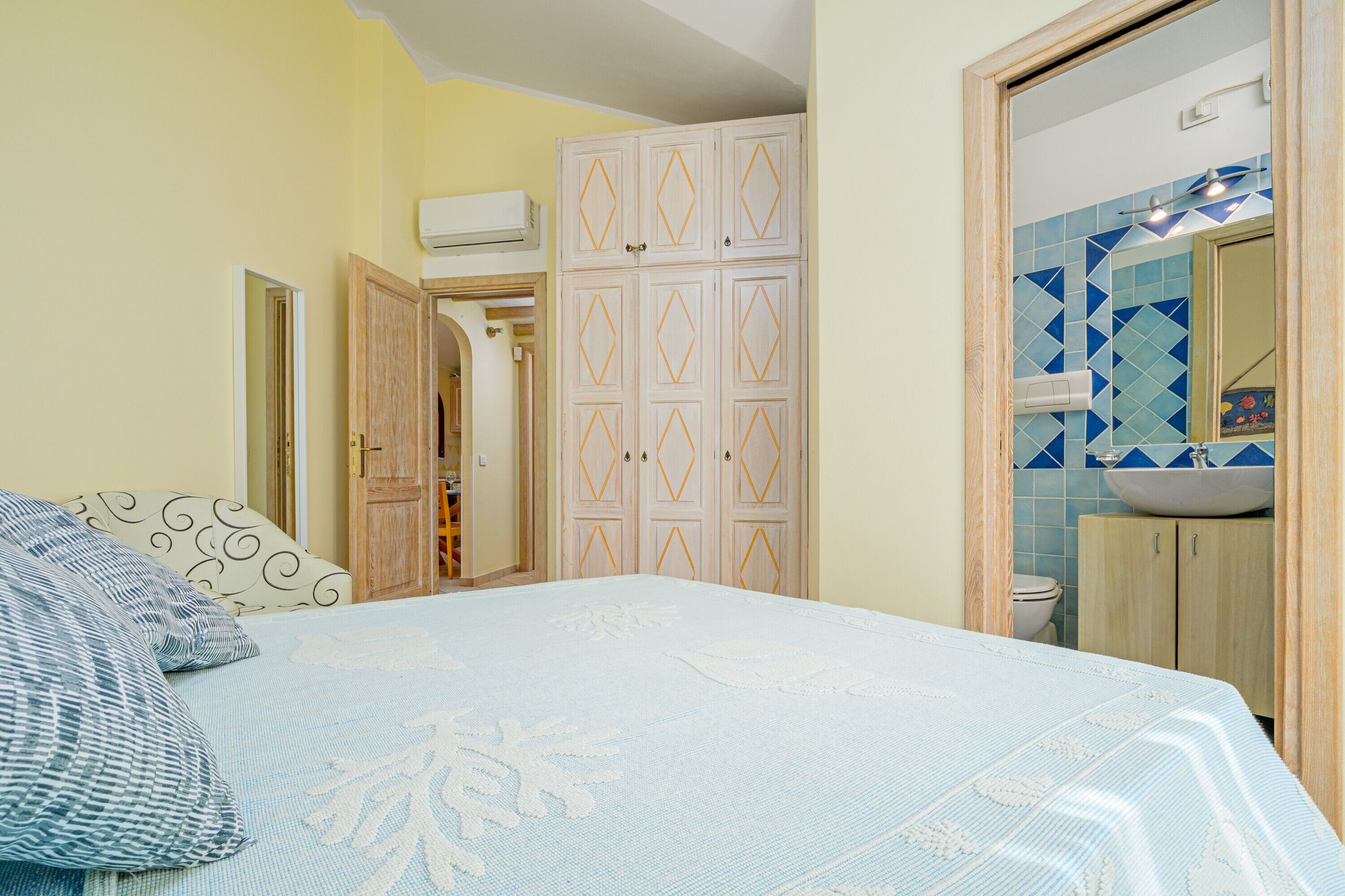 A bedroom with a bed, a wardrobe, a bathroom with a sink and mirror, and a door leading to another room.