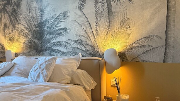 Une chambre avec une grande fresque murale représentant des palmiers tropicaux, un lit avec du linge de lit blanc, une table de chevet avec une lampe et un vase contenant des fleurs séchées.