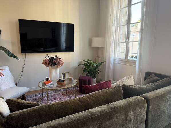 Ein Wohnzimmer mit einer Couch, einem Couchtisch, einem Fernseher, einer Pflanze und einem Fenster mit Vorhängen.