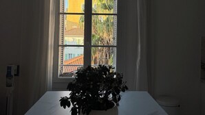 Une pièce avec une grande fenêtre, une table blanche, un objet en bois posé sur la table et une plante en pot.