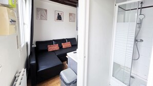 Une pièce de taille modeste, avec un canapé noir, une petite poubelle et un espace douche doté d’une porte en verre.