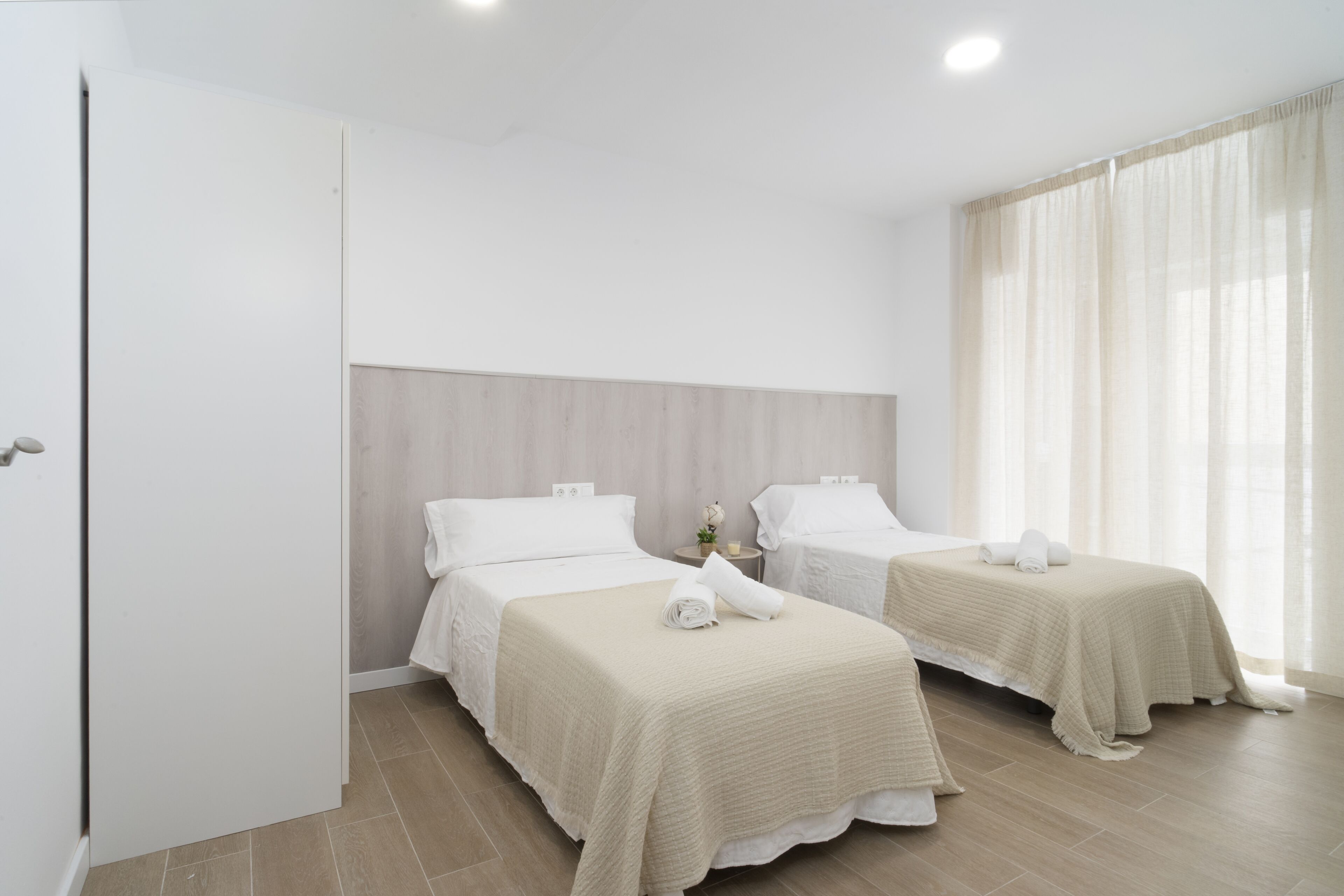 A hotel room with two beds, a wooden headboard, a window with sheer curtains, and a door.