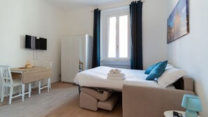 A hotel room with a bed, a desk, a TV, a window with curtains, and a painting on the wall. - GuestHost - Comfortable 30m2 studio, perfect for 2, located on the second floor of a building with lift.The apartment is located in the Ca' Granda - Prato Centenaro area, located in Municipality 9 north of Milan, a well-connected residential area (M5 Ca' (Milan)