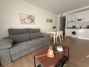 Een moderne woonkamer met een grijze bank, een houten salontafel met een kaars en een boek, een eettafel met stoelen, en een keukenhoek met een wasmachine.
