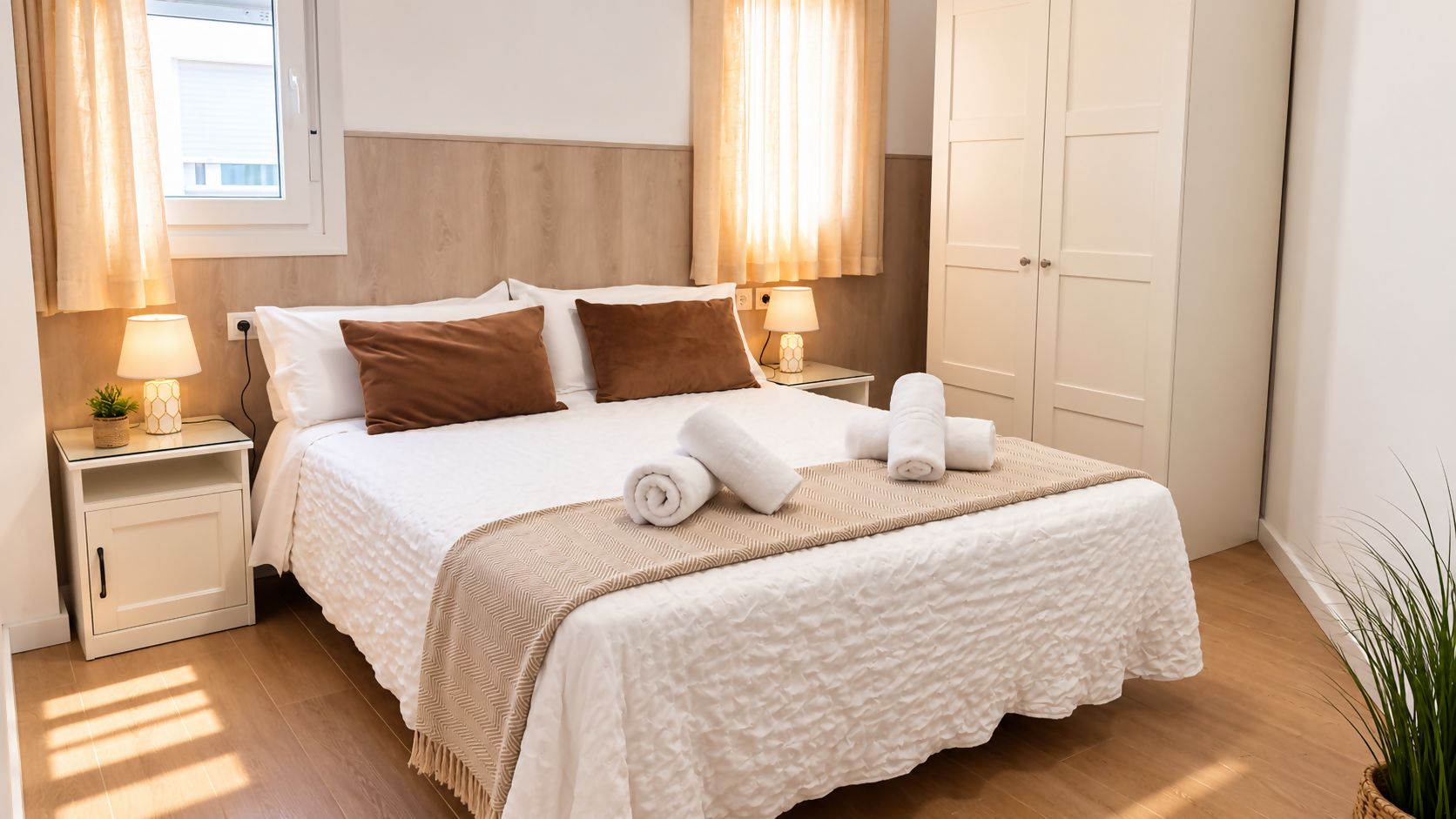 A neatly made bed with a white duvet, brown pillows, and a beige throw blanket. There are two bedside tables with lamps and a potted plant.
