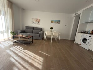 A living room with a sofa, a coffee table, a dining table with chairs, a washing machine, and two wall art pieces. - Apartamento Don José 2Izq (Malaga)