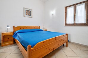 Una cama de madera con colcha azul, una mesita de noche con lámpara, un cuadro enmarcado en la pared y una ventana con cortinas.