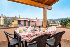 Un patio techado con una mesa preparada para cuatro personas, rodeado de sillas de mimbre, con vistas a una zona residencial con edificios coloridos y una montaña al fondo.
