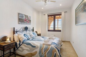 A bedroom with a bed, a nightstand, a ceiling fan, a window with curtains, and a painting on the wall.