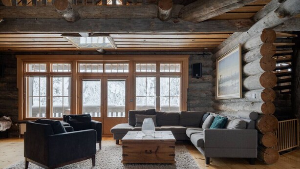 L’intérieur d’une cabane en rondins, avec un plancher en bois, un ensemble de canapés, une table basse et de grandes fenêtres donnant sur des arbres couverts de neige.