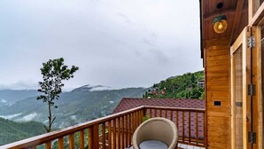 Un balcon en bois offrant une vue sur un paysage montagneux, une chaise en osier et une petite table avec une bouilloire et une tasse.
