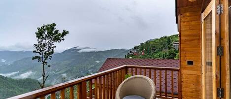 A wooden balcony with a view of a mountainous landscape, a wicker chair, and a small table with a kettle and cup.