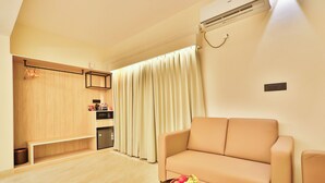 A living room with a sofa, a small table with fruit, a kitchen area with a microwave, and a window with curtains.