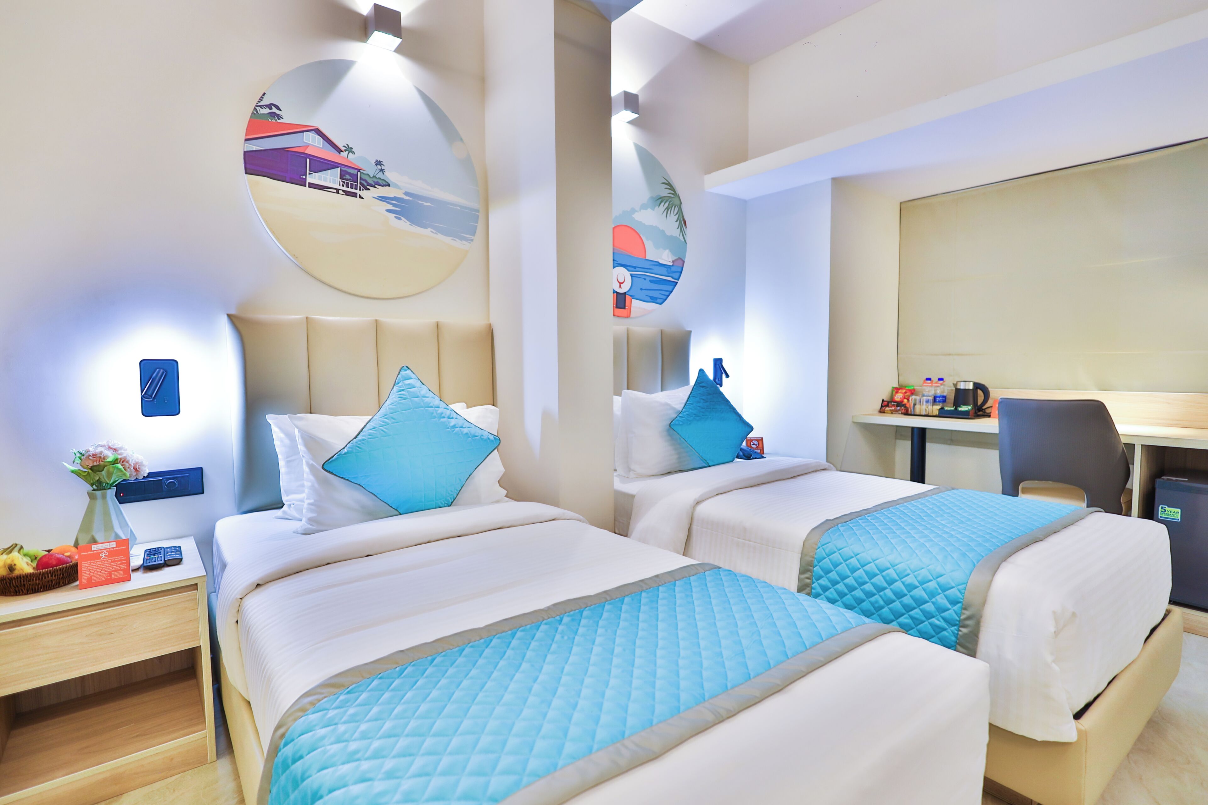 A hotel room with two beds, a desk, a chair, and a small table with a basket of fruit.