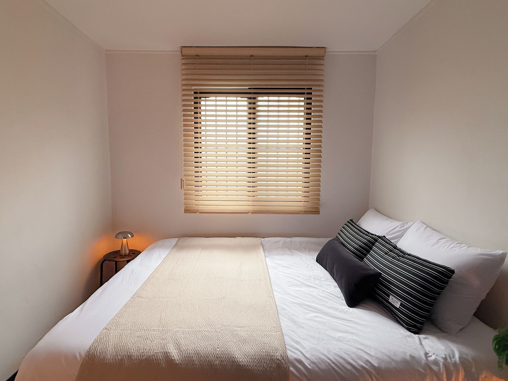 Ein Schlafzimmer mit einem Bett, einem Fenster mit Jalousien, einer Nachttischlampe und einer Pflanze.
