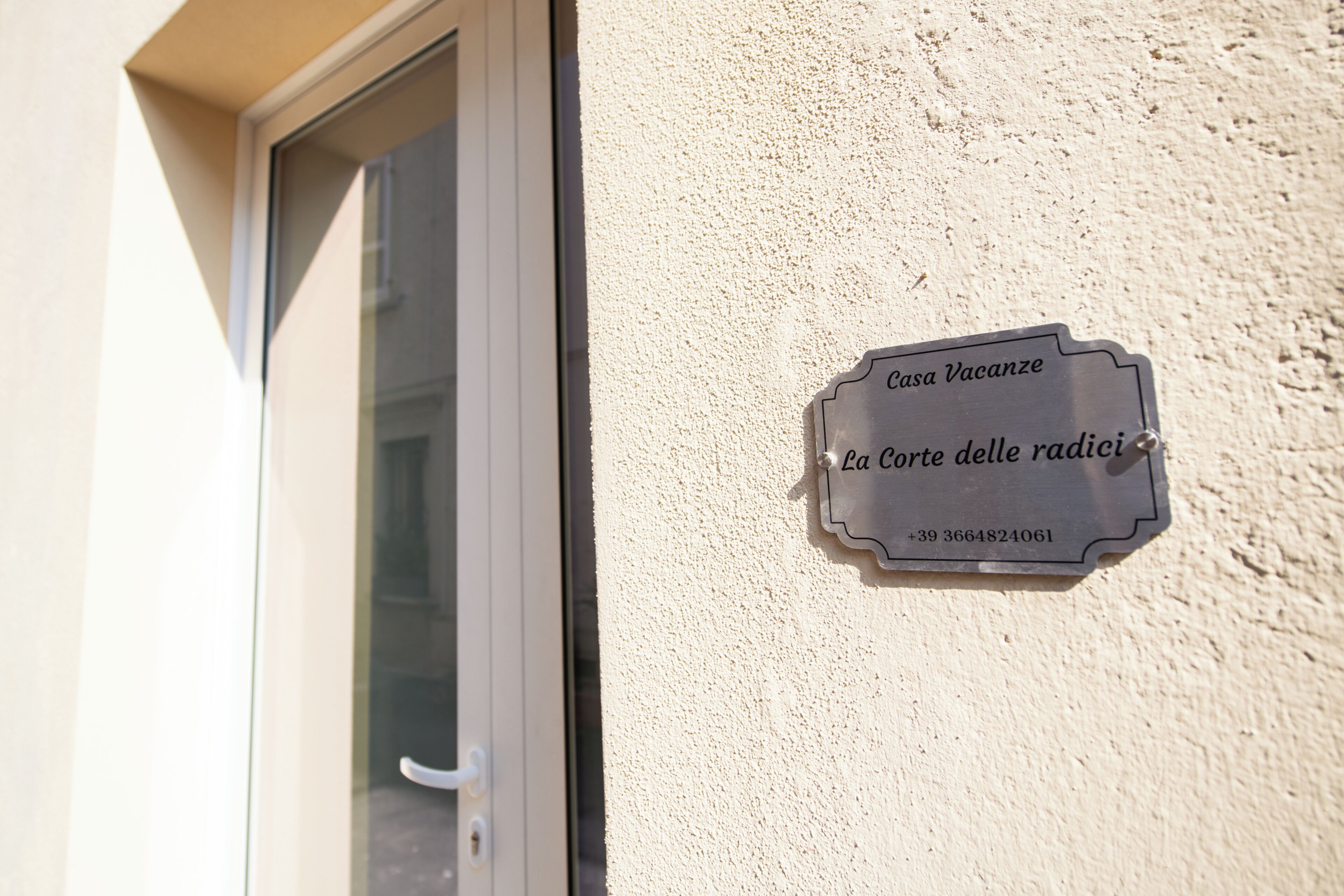 A metal plaque with the text 'Casa Vacanze La Corte delle radici' and a phone number '+39 3664824061' affixed to a textured wall.