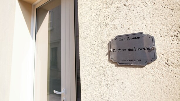 A metal plaque with the text 'Casa Vacanze La Corte delle radici' and a phone number '+39 3664824061' affixed to a textured wall.