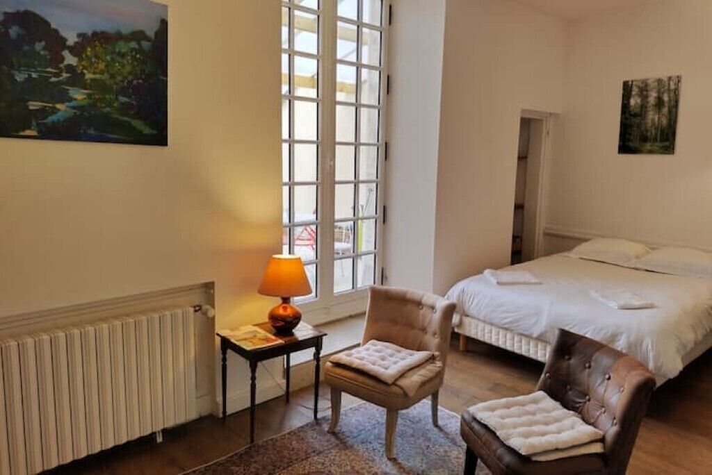 Quarto com cama, duas cadeiras, mesa com abajur e um quadro na parede.