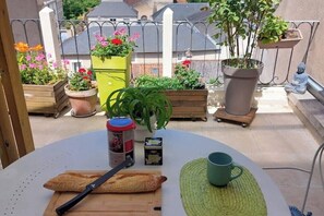 A round table with a green mat, a green mug, a red container, and a baguette on a balcony with potted plants and a statue. - Downtown studio apartment with a spacious private terrace—quiet and bright (Poitiers)