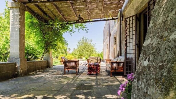 A covered outdoor seating area with stone flooring, wooden furniture, and a stone wall.