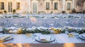 An outdoor dining area with tables set for a meal, clear chairs, and a building with arched windows in the background.