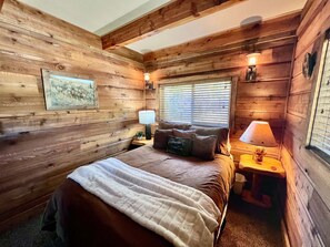 Une chambre à coucher dans un chalet en bois, avec un lit, deux lampes de chevet, une fenêtre avec des stores et un tableau encadré au mur.