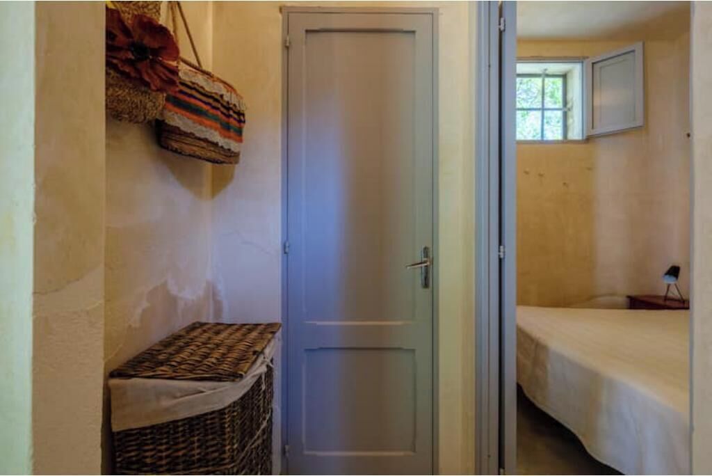A narrow hallway with a door, a small wicker basket, and a bed with a white sheet in the adjacent room.