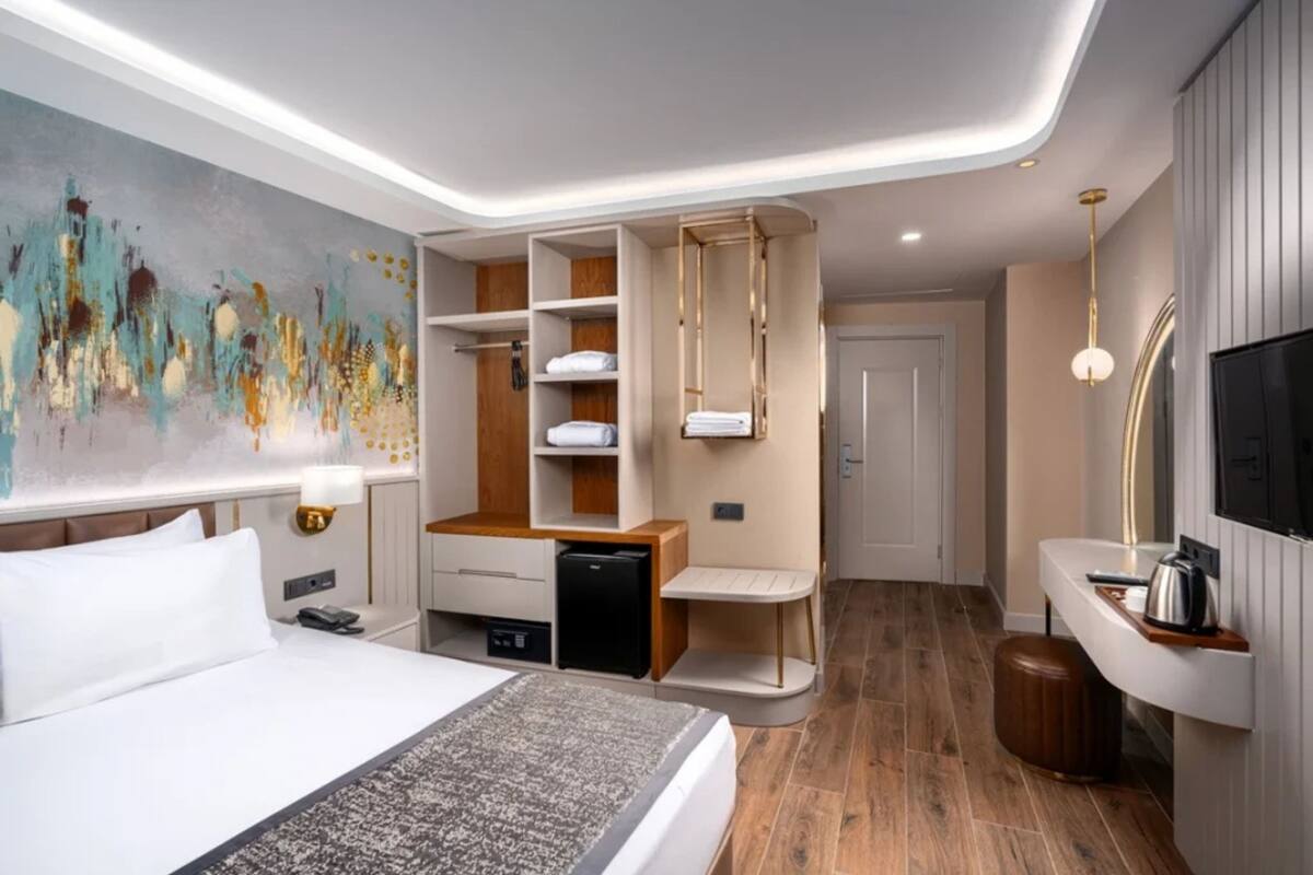 A modern hotel room with a bed, a desk, a wardrobe, a television, and a painting on the wall.