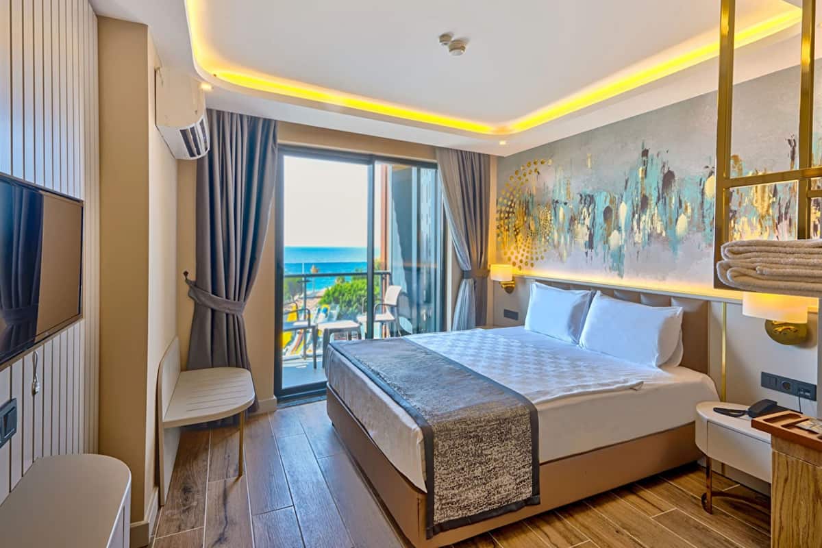 A hotel room with a large bed, a TV, a desk, a chair, and a balcony with a view of the sea.