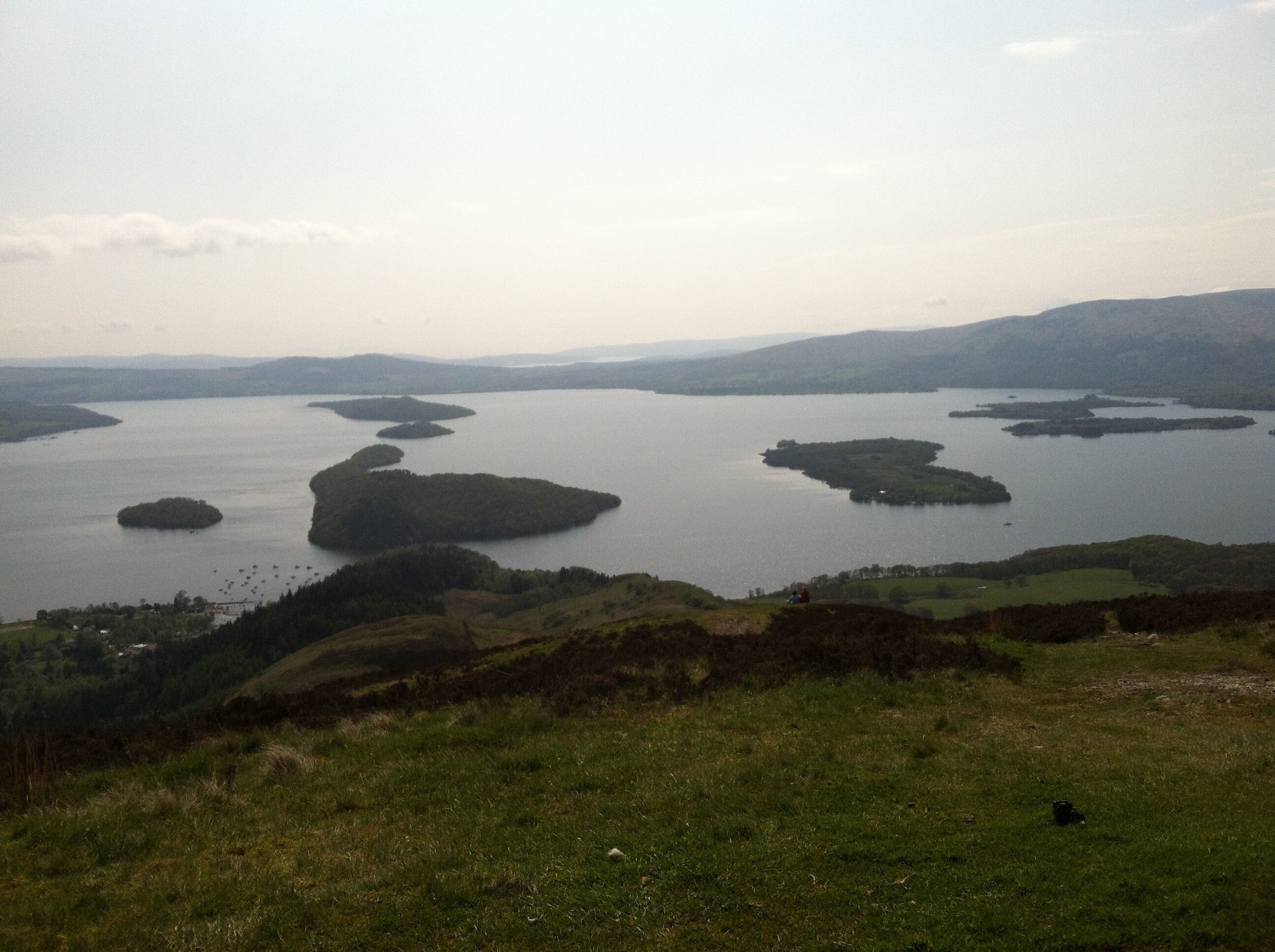 A panoramic view of a lake with multiple islands, surrounded by hills and a grassy foreground.