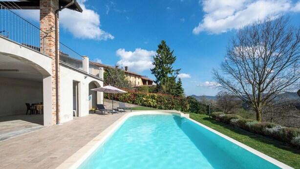 Una piscina con agua azul cristalina, rodeada por un patio con mobiliario exterior, una columna de ladrillo y vistas al paisaje.