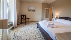 Habitación de hotel con cama, una silla, radiador, una mesa de vidrio y un cuadro en la pared.