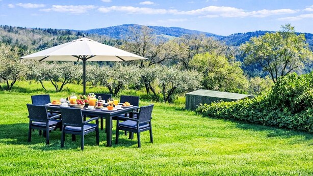 Een terras met een ontbijttafel, omringd door stoelen en een parasol, met uitzicht op een bergachtig landschap.