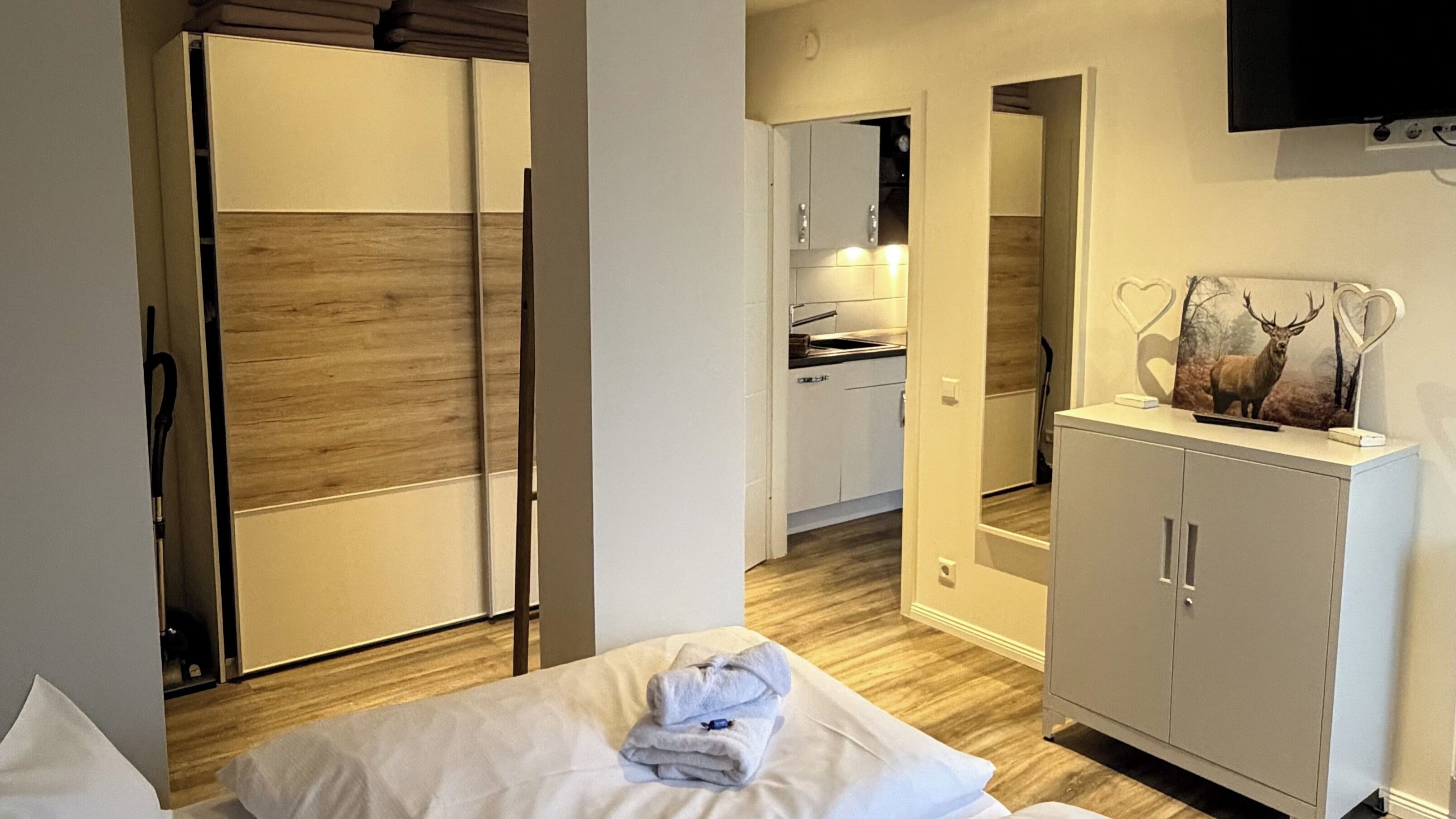 A hotel room with a wooden floor, a bed with white linens, a wardrobe, a mirror, and a television mounted on the wall.