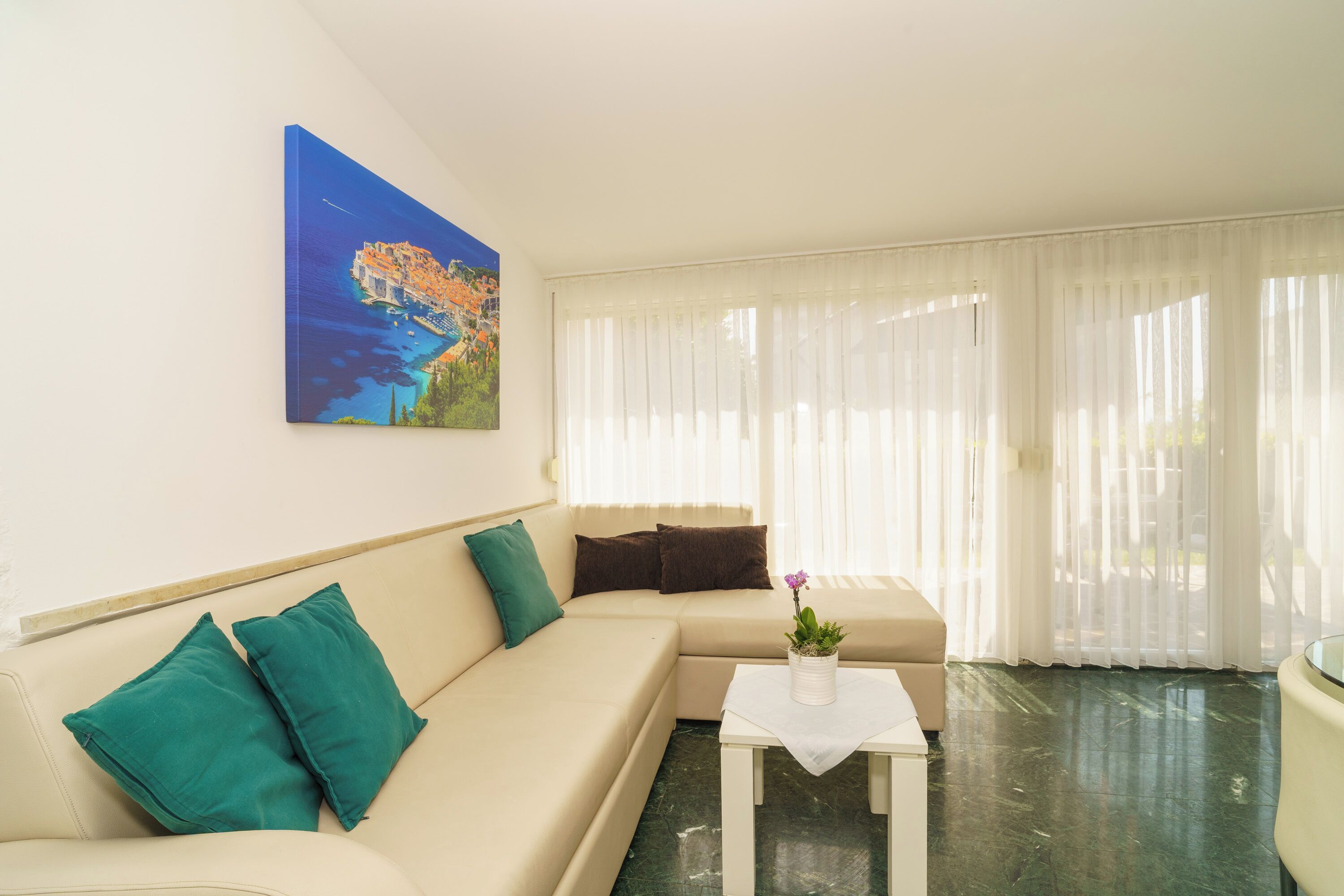 Sala de estar com um sofá bege, almofadas verde-água, uma pequena mesa branca com uma planta em vaso e um quadro na parede.