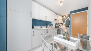 Een moderne keuken met witte kasten, een blauwe accentmuur en een eethoek met een glazen tafel en stoelen.