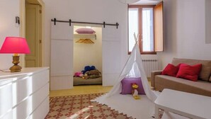 Ett rum med en vit byrå, en lampa, en soffa med röda kuddar, en vit stol, ett fönster med utsikt och en dörr som leder till ett annat rum.