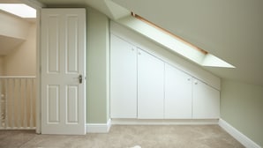 Ein Zimmer mit schräg verlaufender Decke, einer weißen Tür und einem Schrank mit mehreren Türen.