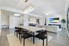 A modern living room with a dining area, a kitchen, and a large TV screen. - Quattro at Naples Square (Naples)